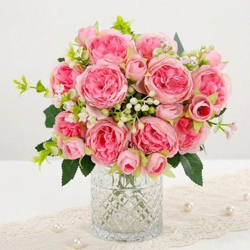 Pink faux peony bouquet with layered petals and green leaves in a textured silver vase.