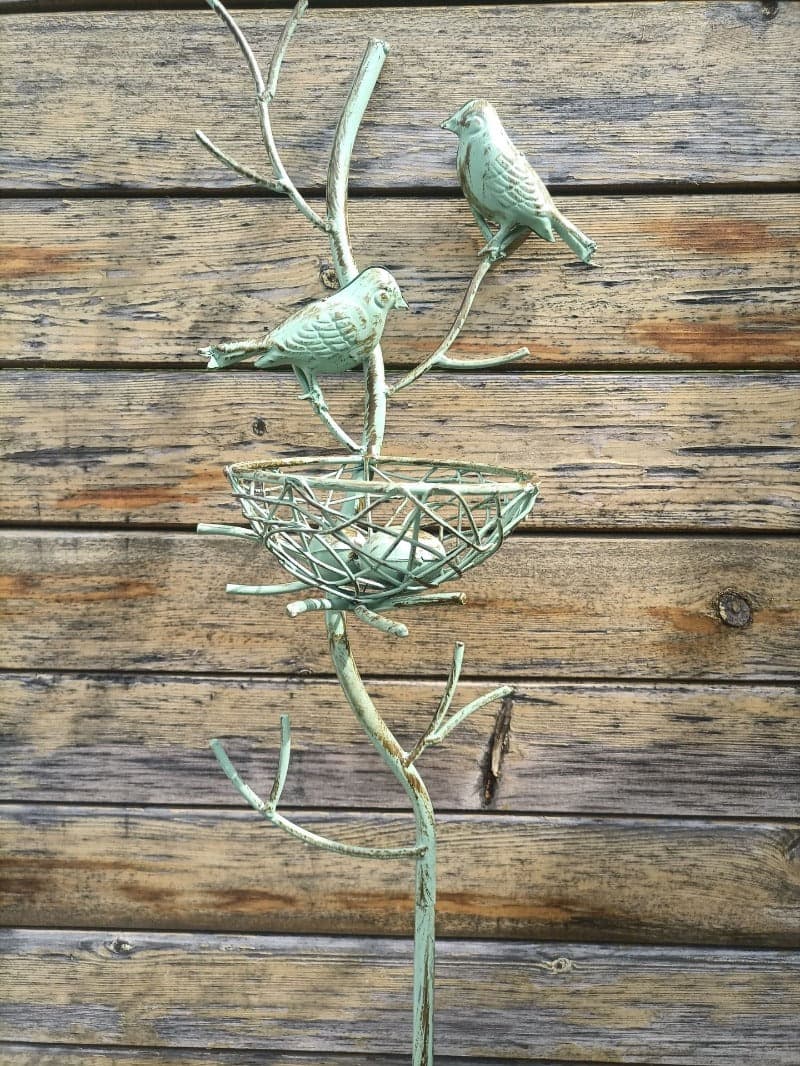 Green metal garden trellis stake with two birds perched above a wire nest basket