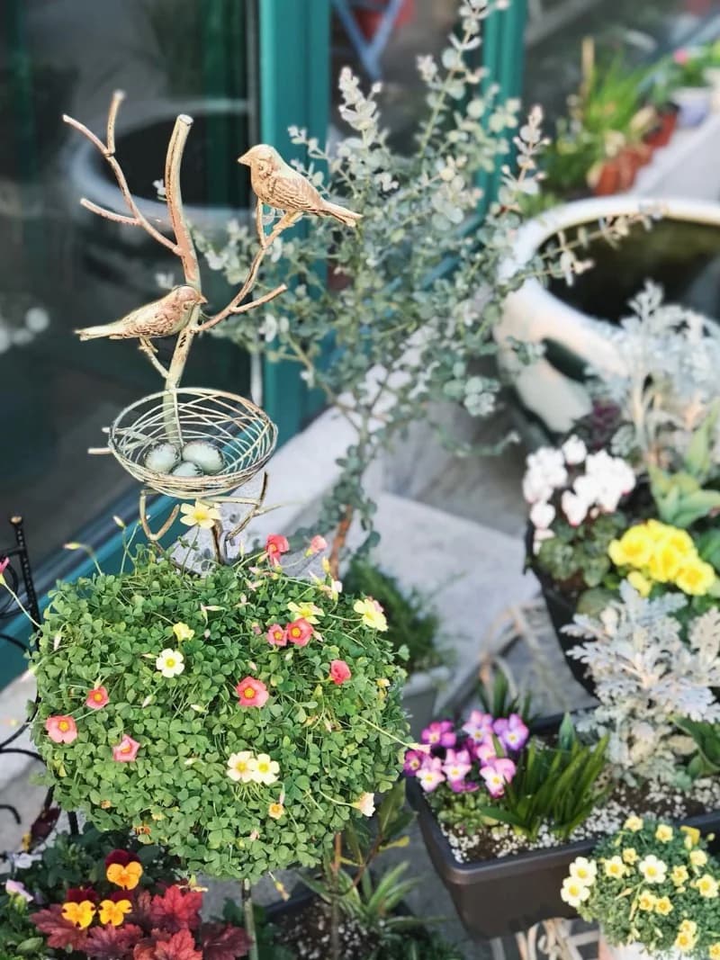 Green metal bird trellis stake with wire nest basket and perched birds among potted flowers