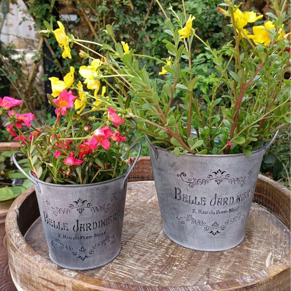 Two metal planters with flowers on a wooden surface outdoors.