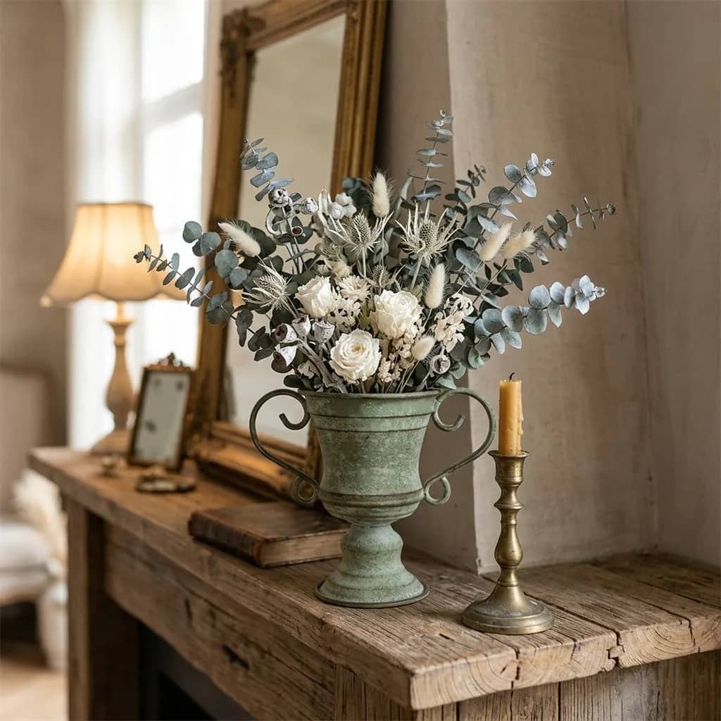 Decorative floral arrangement in a vintage urn on a wooden table with a candle and mirror in the background.