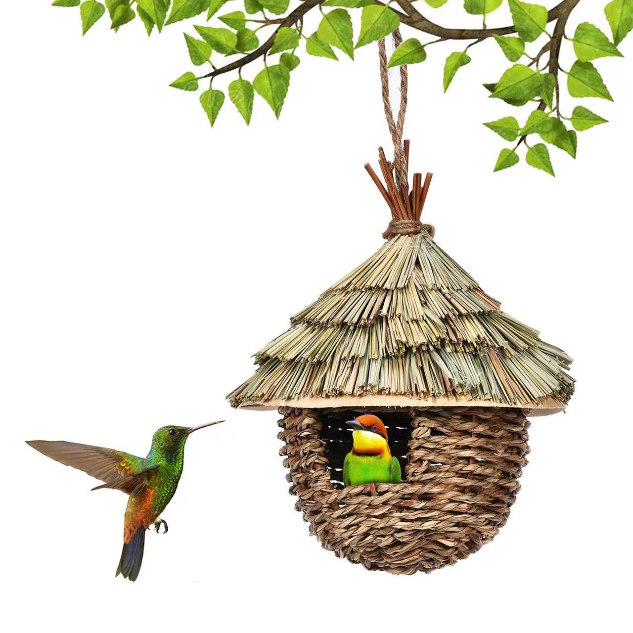 Woven birdhouse with a thatched roof, hanging from a branch with leaves, on a white background.