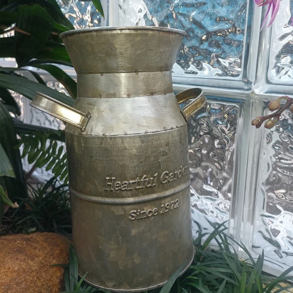 Mælkedåse i kobber med sidehåndtag og stemplet logo, placeret blandt planter og græs.