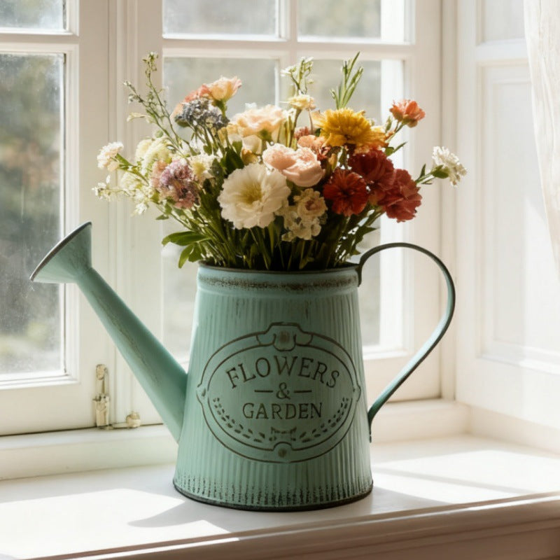 Teal metal watering can vase with 'Flowers & Garden' emblem, filled with multicolor cut flowers.