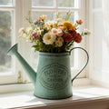Teal metal watering can vase with 'Flowers & Garden' emblem, filled with multicolor cut flowers.