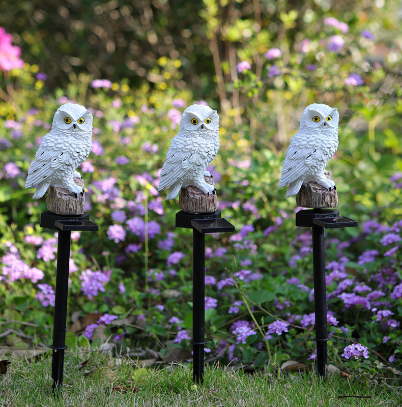 Three white owl solar garden stake lights with glowing bases among flowers
