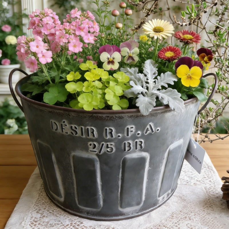 Galvaniseret metalplantespand med håndtag og præget skrift, fyldt med blomster