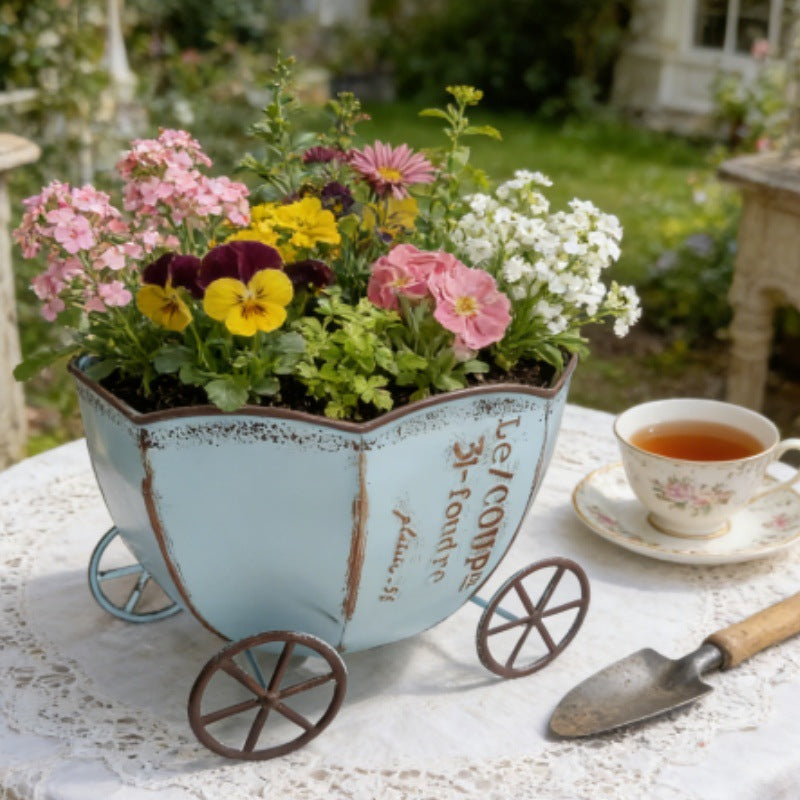 Vintage metalplantekasse med blågrå finish og blomstermix på fire hjul