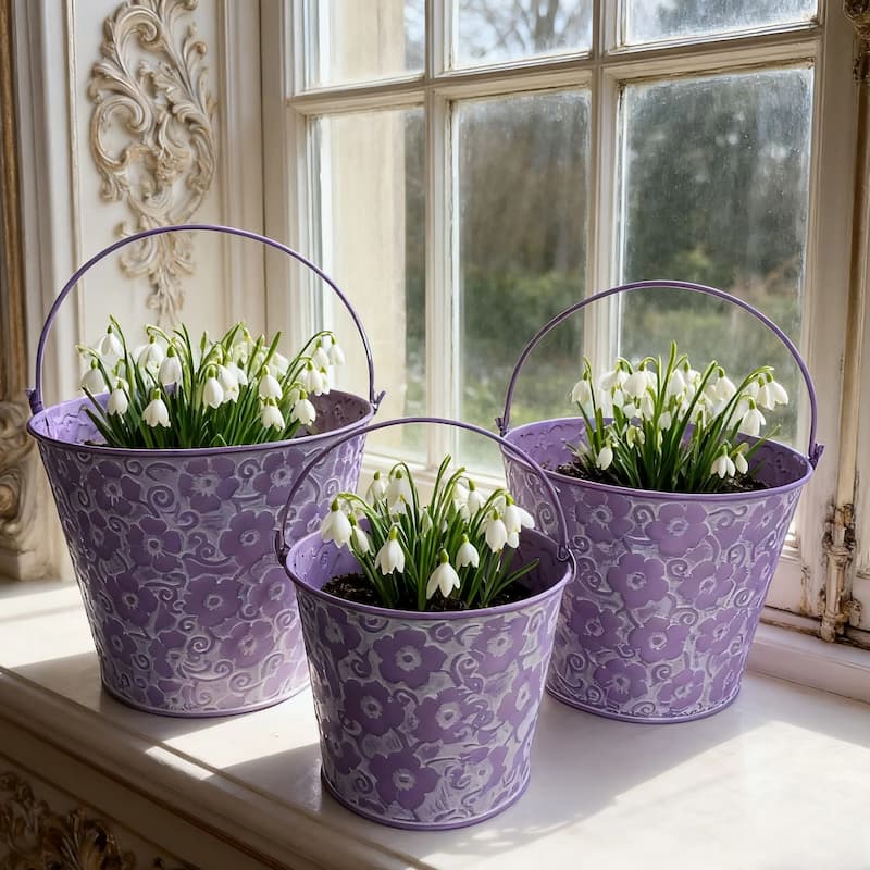 Sæt med 3 lilla prægede metalplantekasser med buede håndtag, fyldt med blomster.