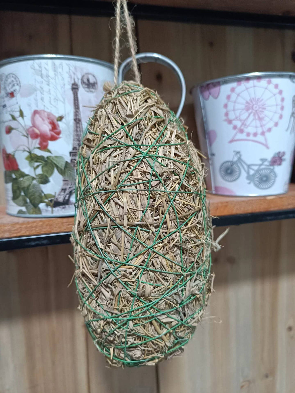 Hanging bird nest made of straw and twine on a wooden shelf with decorative items.