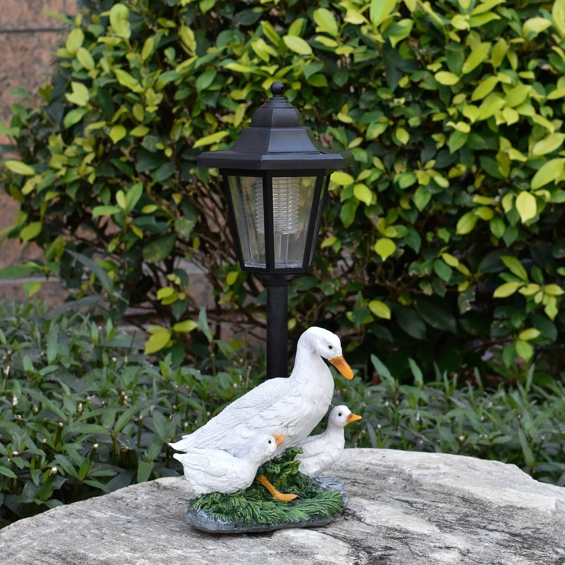 Decorative duck family sculpture with a lamp post on a stone surface in front of green foliage.
