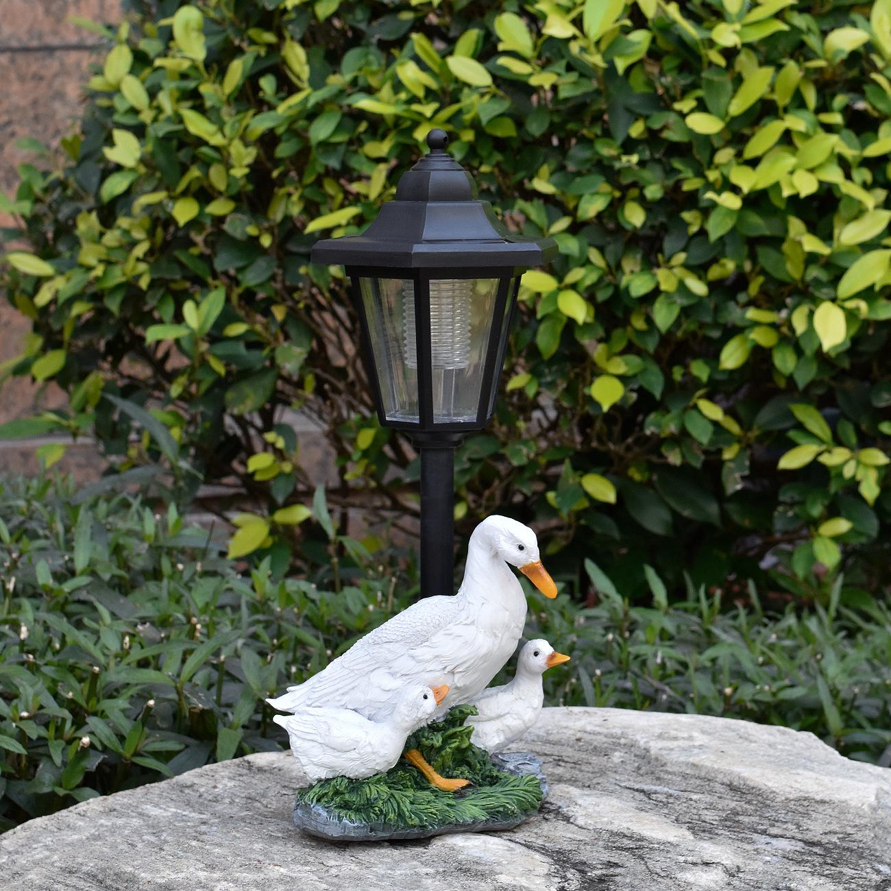 Decorative duck family sculpture with a lamp post on a stone surface in front of green foliage.
