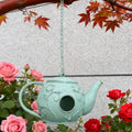 Teal birdhouse shaped like a teapot hanging among pink and red roses.