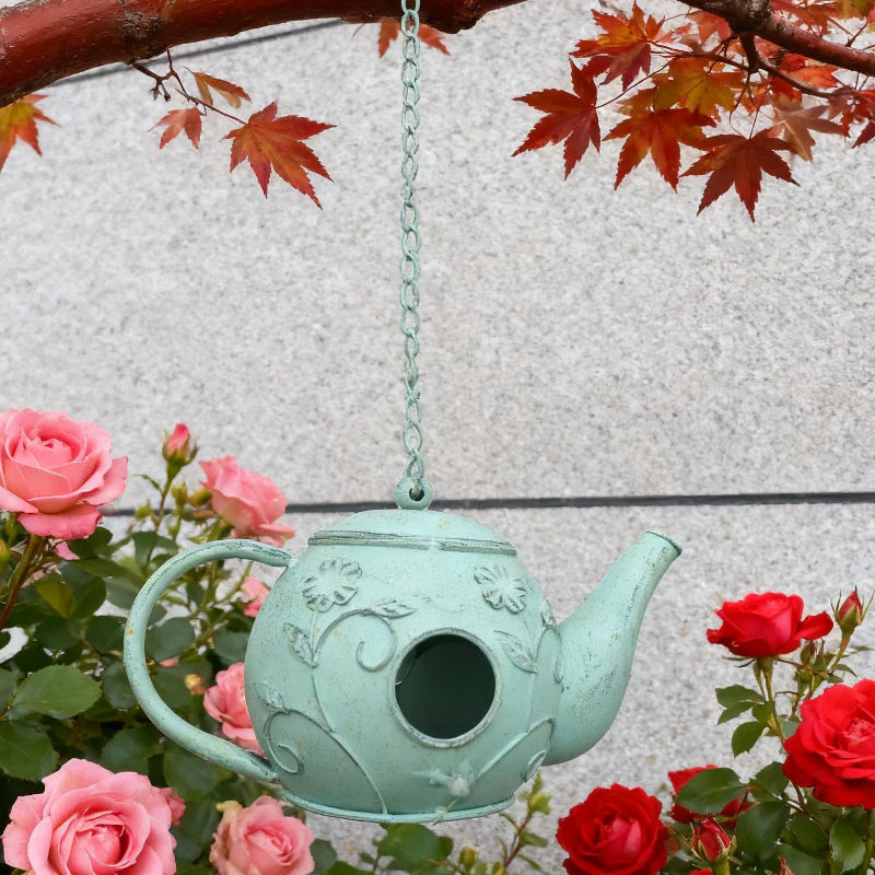 Teal birdhouse shaped like a teapot hanging among pink and red roses.