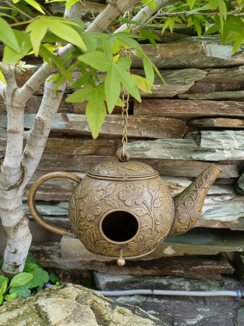 Decorative teapot birdhouse hanging against a stone wall with greenery.