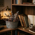 Decorative arrangement of pinecones and branches in a metal container on a wooden shelf with books and a magnifying glass.