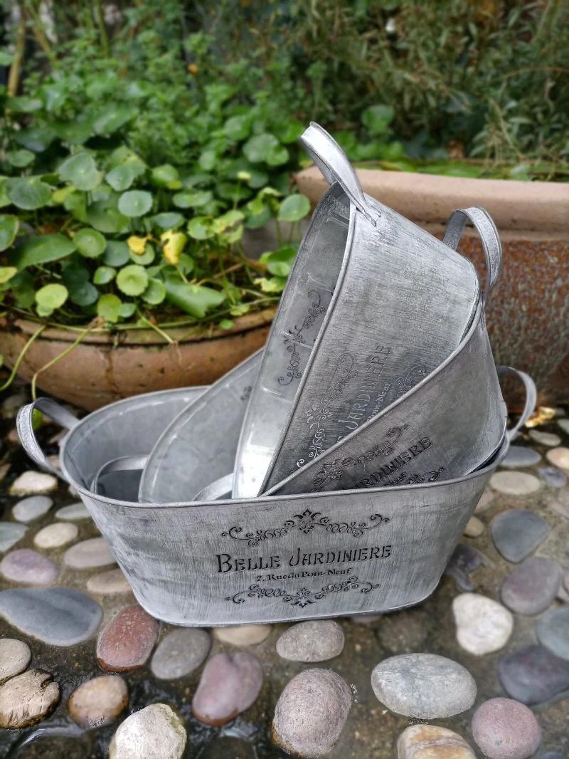 Three metal planters with 'Belle Jardinière' branding on a stone surface with greenery in the background.