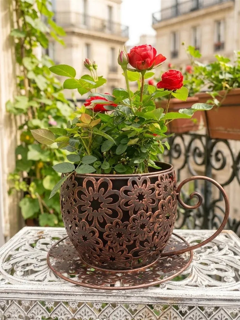 Decorative metal planter shaped like a teacup with red flowers on a balcony.Vintage Floral Cutout Metal Teacup Planter – Rustic Iron Cachepot with Saucer Base & Scrolled Handle (11" Wide)